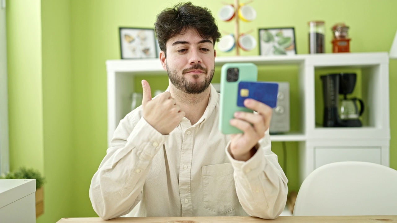 年轻的西班牙裔男子坐在餐桌上用智能手机和信用卡购物视频素材