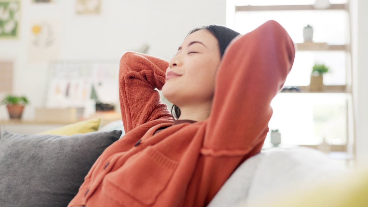 亚洲女性，周末在家里或客厅的沙发上快乐或放松，休息日或禅心健康。微笑，人或手放在头后的沙发上，平静或自我照顾的心态从休息视频素材