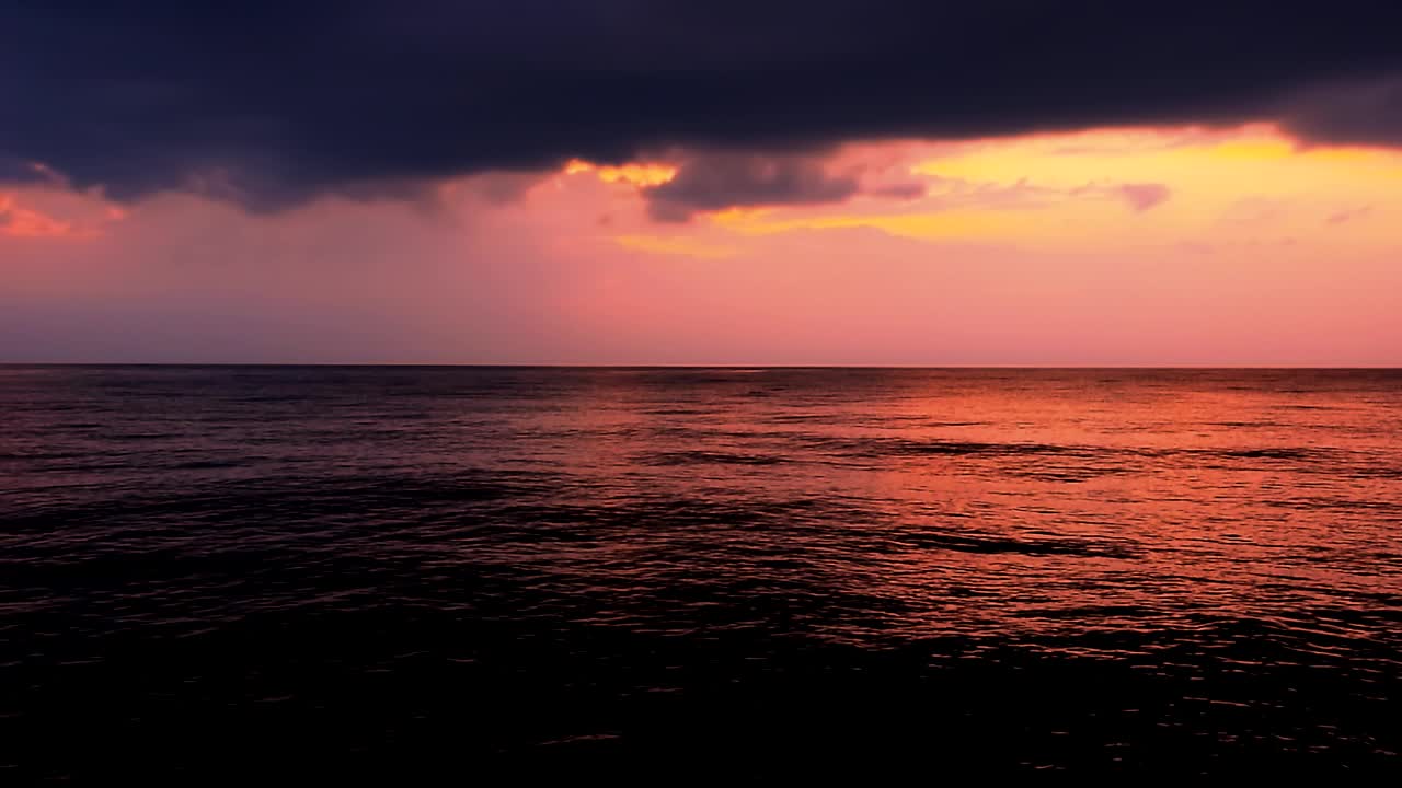 在紫色的夕阳背景上，海浪反射出红色的晚霞。美丽的阳光在粉红色和紫色的天空与云，平静的场景由地平线傍晚由阳光明媚的海滩，中景，慢动作，高清。ProRes 422 HQ。视频素材