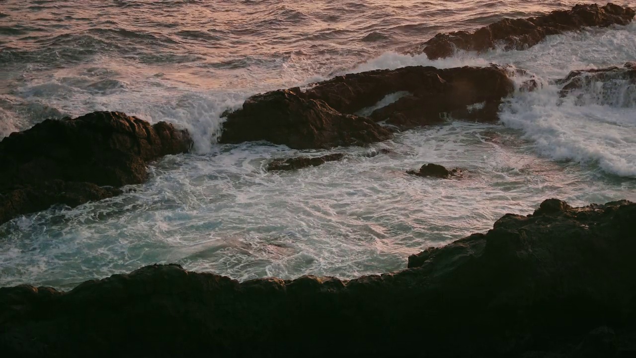 在温暖的夕阳下，海浪拍打着岩石海滩。特写:狂暴的暴风雨泡沫，水溅在火山岸边。视频素材