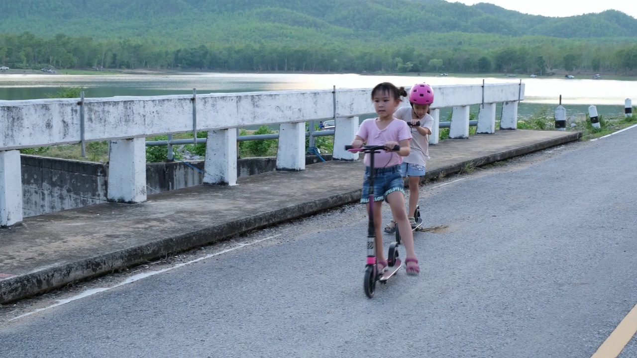 两个可爱的小女孩在夏季公园的户外道路上骑着滑板车视频素材