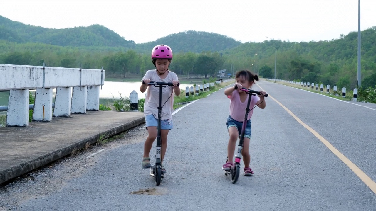 两个可爱的小女孩在夏季公园的户外道路上骑着滑板车视频素材