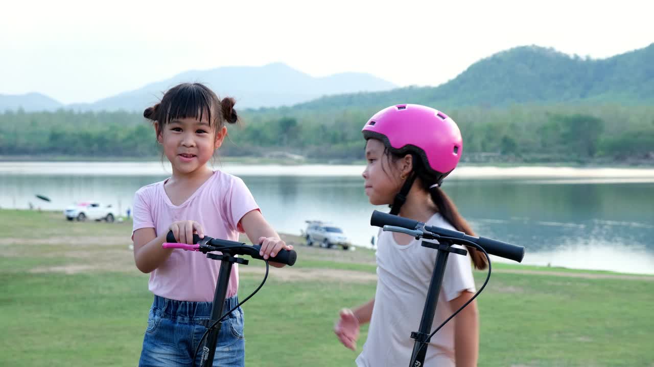 两个可爱的小女孩在夏天的花园里微笑着摆姿势。在公园里骑滑板车的快乐小孩。儿童的健康运动和户外活动。视频素材