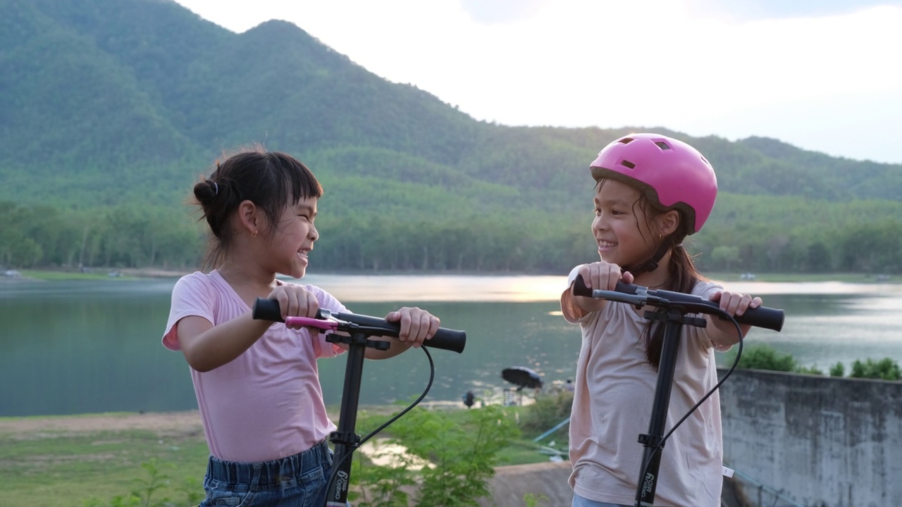 两个可爱的小女孩在夏天的花园里微笑着摆姿势。在公园里骑滑板车的快乐小孩。儿童的健康运动和户外活动。视频素材