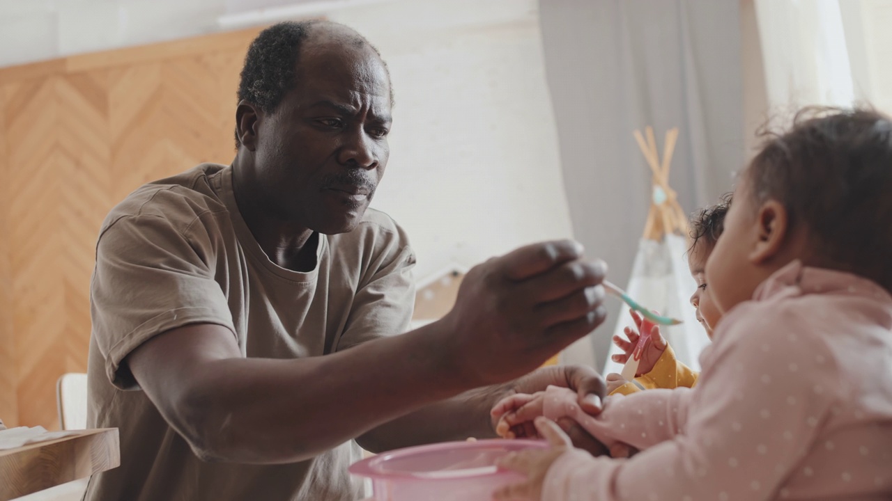 非裔美国父亲给坐在高脚椅上的两个婴儿喂食视频素材