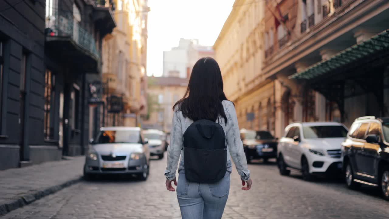 镜头跟随一位微笑的年轻女子走在城市街道上。一个美丽的旅游女孩背着背包，在城市的背景下微笑着，环顾四周，欣赏着城市的建筑视频素材