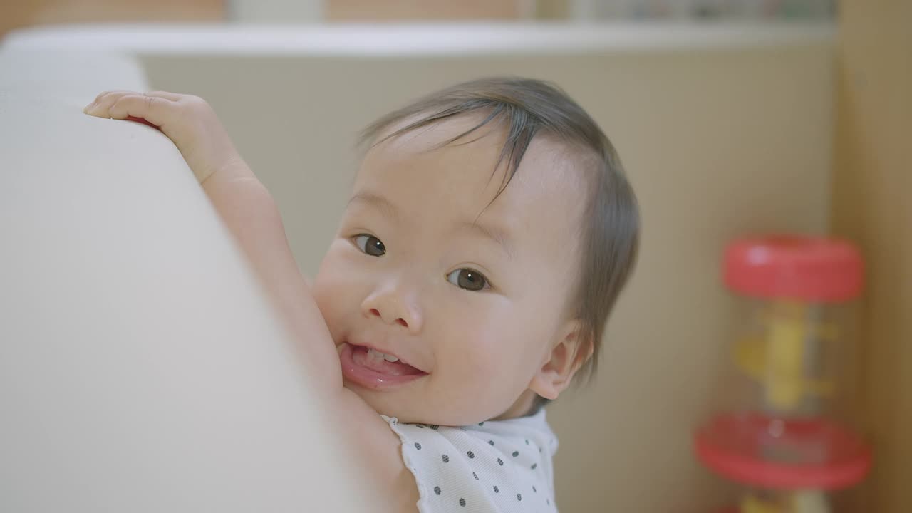 可爱的小女婴的脸，带着兴趣和惊讶地抓着镜头，爸爸在家里的客厅里拍摄蹒跚学步的婴儿视频素材