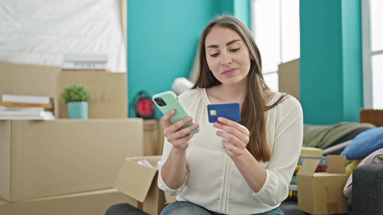 年轻漂亮的西班牙女人用智能手机和信用卡坐在新家的沙发上购物视频素材