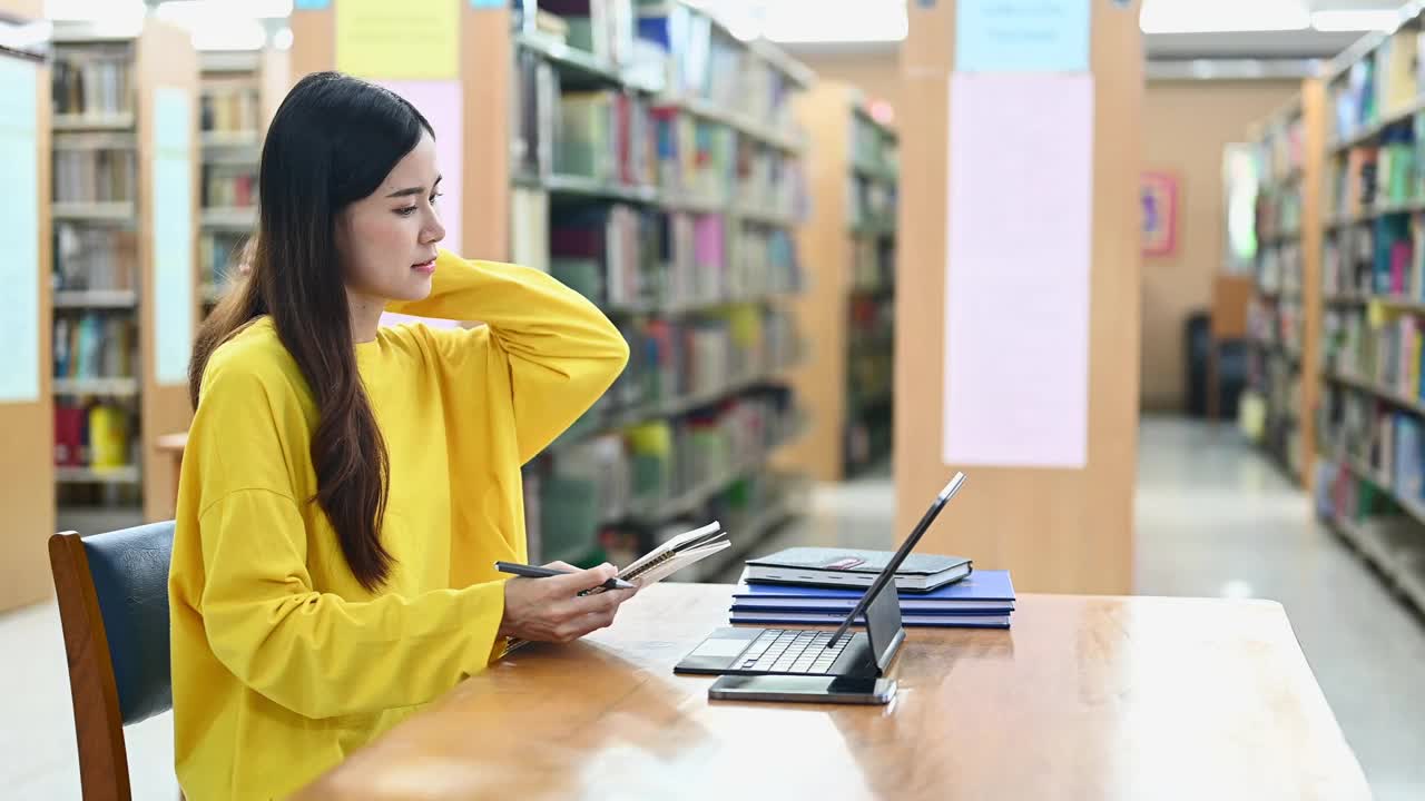 微笑的亚洲女学生坐在大学图书馆做作业，在笔记本电脑上搜索信息。视频素材