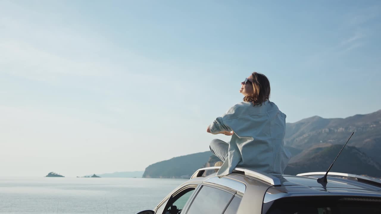 一个年轻的白人妇女坐在她停在海边的车顶上视频素材