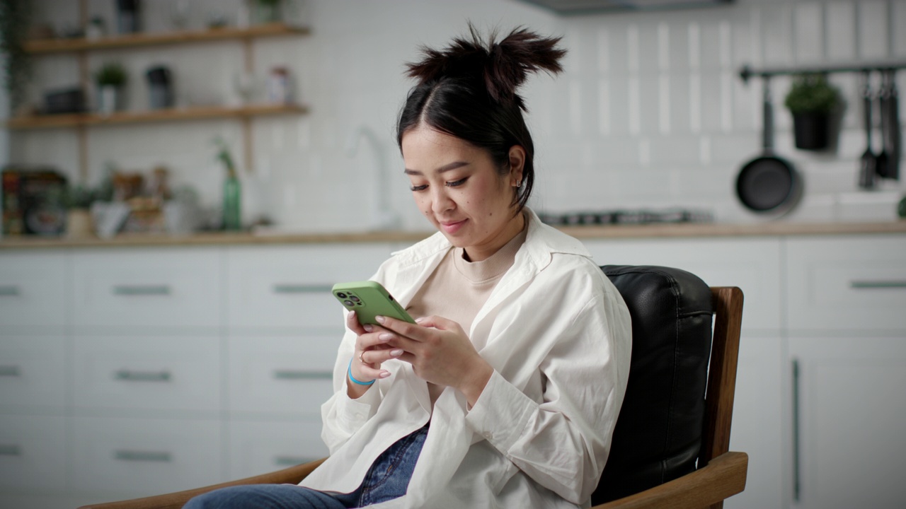 女人用智能手机发短信。亚洲韩国女性坐在家里的扶手椅上微笑着与朋友在线聊天。视频素材