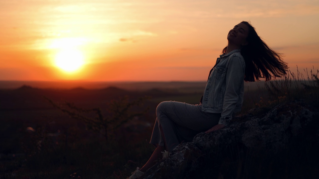 一位快乐的长发女子欣赏着日落时晚霞的清新。在夕阳的背景下，女性的侧面轮廓与松散的头发。风吹过头发视频素材