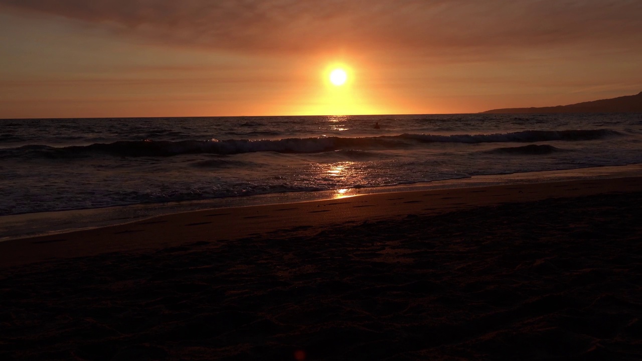 一个人影在海浪中游泳，身后是灿烂的橙色夕阳。视频下载