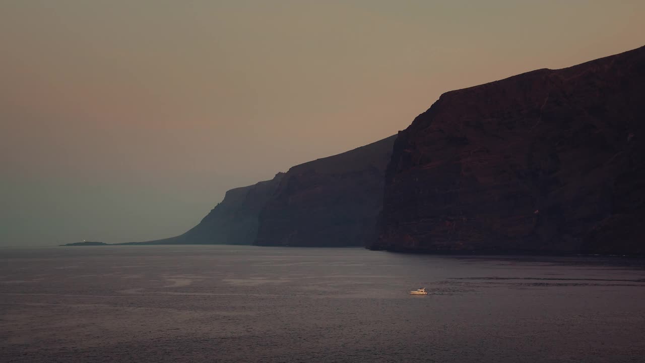 崎岖的海岸线和孤独的小船在黑夜的海洋中航行。海军蓝的海水，表面光滑如玻璃。视频素材