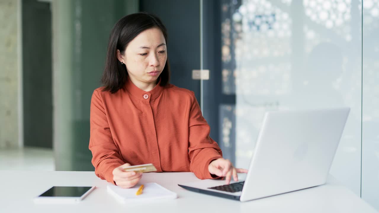 一位亚洲女商人坐在现代办公室的工作场所，通过在笔记本电脑上输入信用卡号码来检测欺诈视频素材