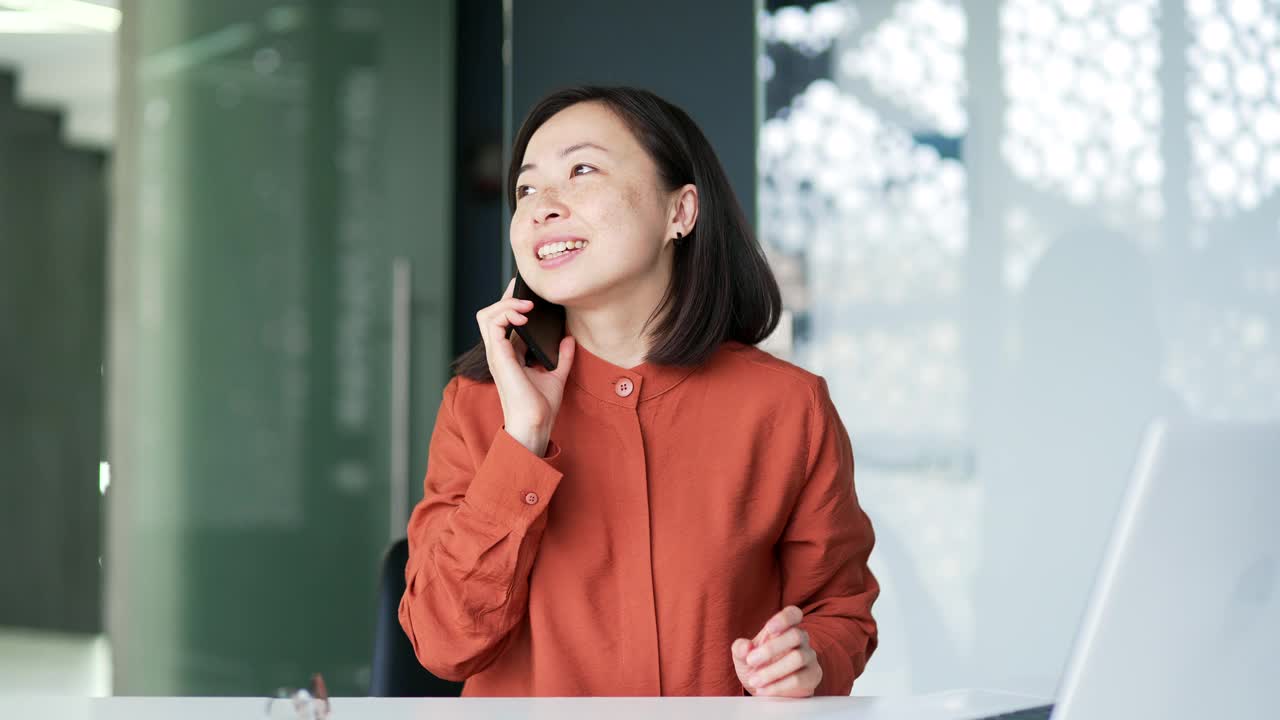 快乐友好的亚洲女性坐在现代办公室的办公桌前，一边用智能手机聊天。视频素材