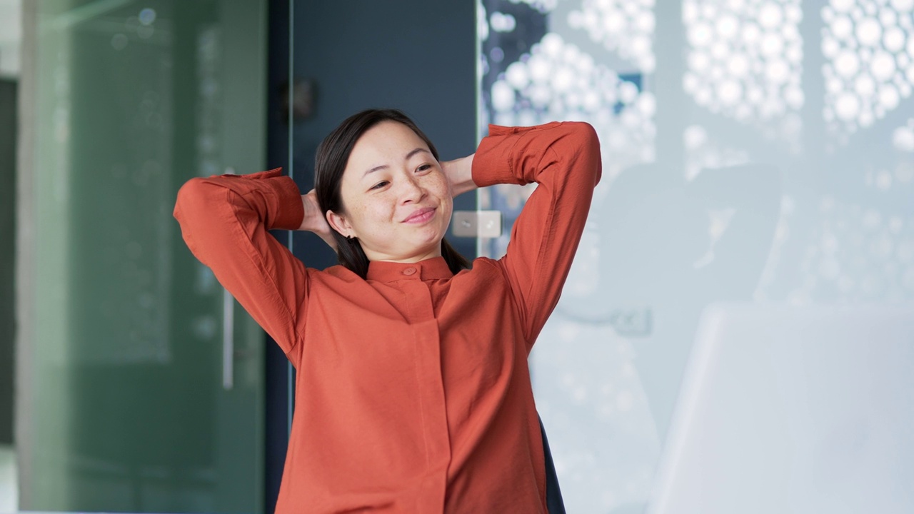 一位面带微笑的亚洲女商人在一台笔记本电脑上完成了工作，坐在现代办公室的工作场所，双手放在脑后放松视频素材