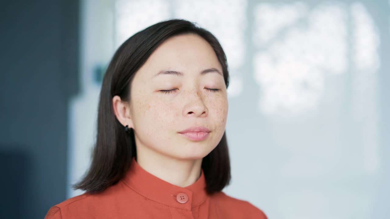 一个亚洲女人闭着眼睛在现代办公室里放松的特写。一个平静快乐的女性休息的肖像视频素材