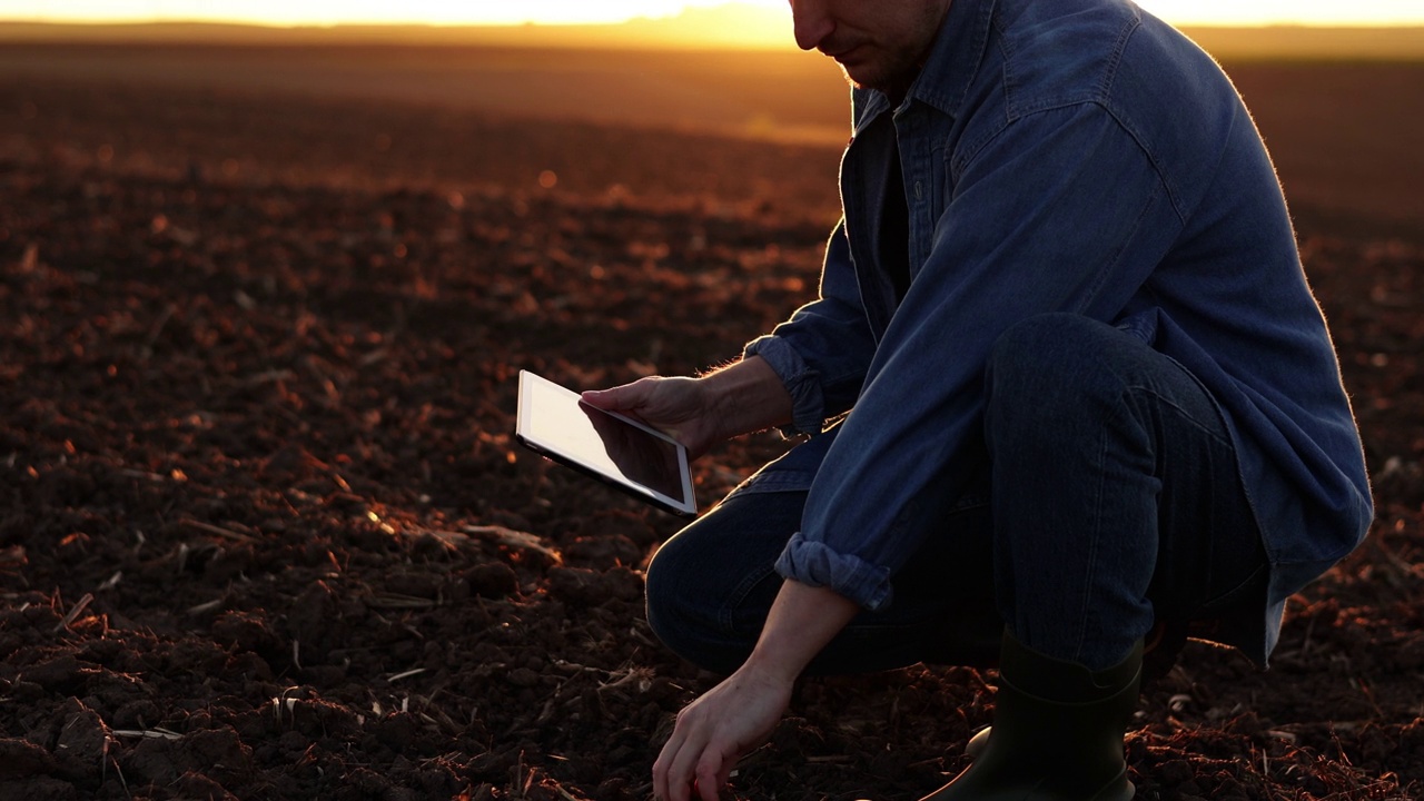 日落时分，一名男性农民在犁过的农田上播种前，触摸土壤，用数码平板检查和测试耕地。智能农业技术和有机农业。视频素材