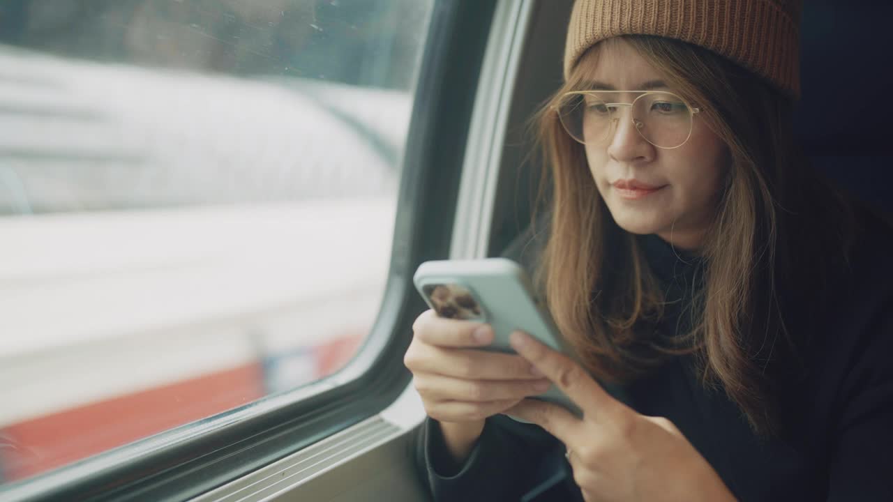 亚洲年轻女性自由职业者游客坐在公共交通列车的窗口边玩智能手机。视频素材