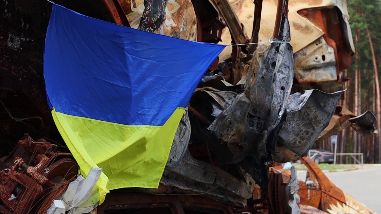 在废墟和被毁汽车生锈的金属背景下的乌克兰国旗。蓝黄旗在阳光下迎风飘扬视频下载