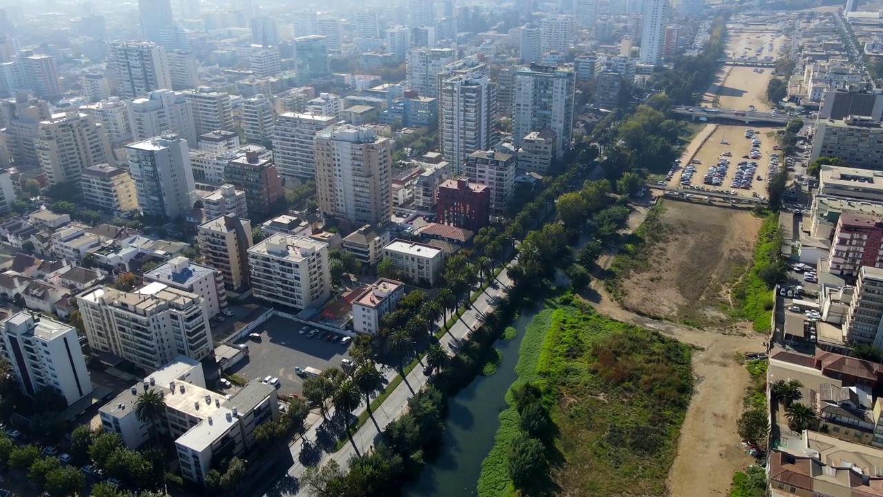 航拍无人机向前飞过河流，旁边的道路两旁是树木，旁边是市中心的大型建筑物视频素材