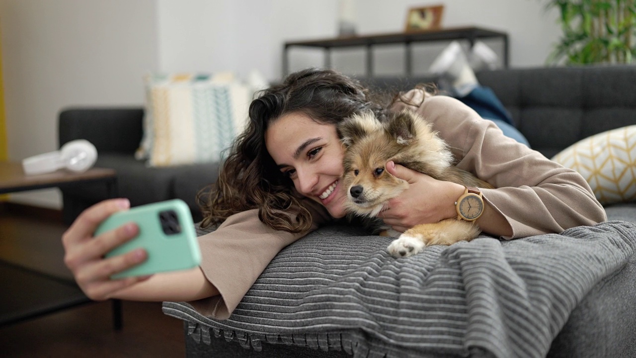 年轻的西班牙女子带着狗躺在家里的沙发上用智能手机自拍视频素材