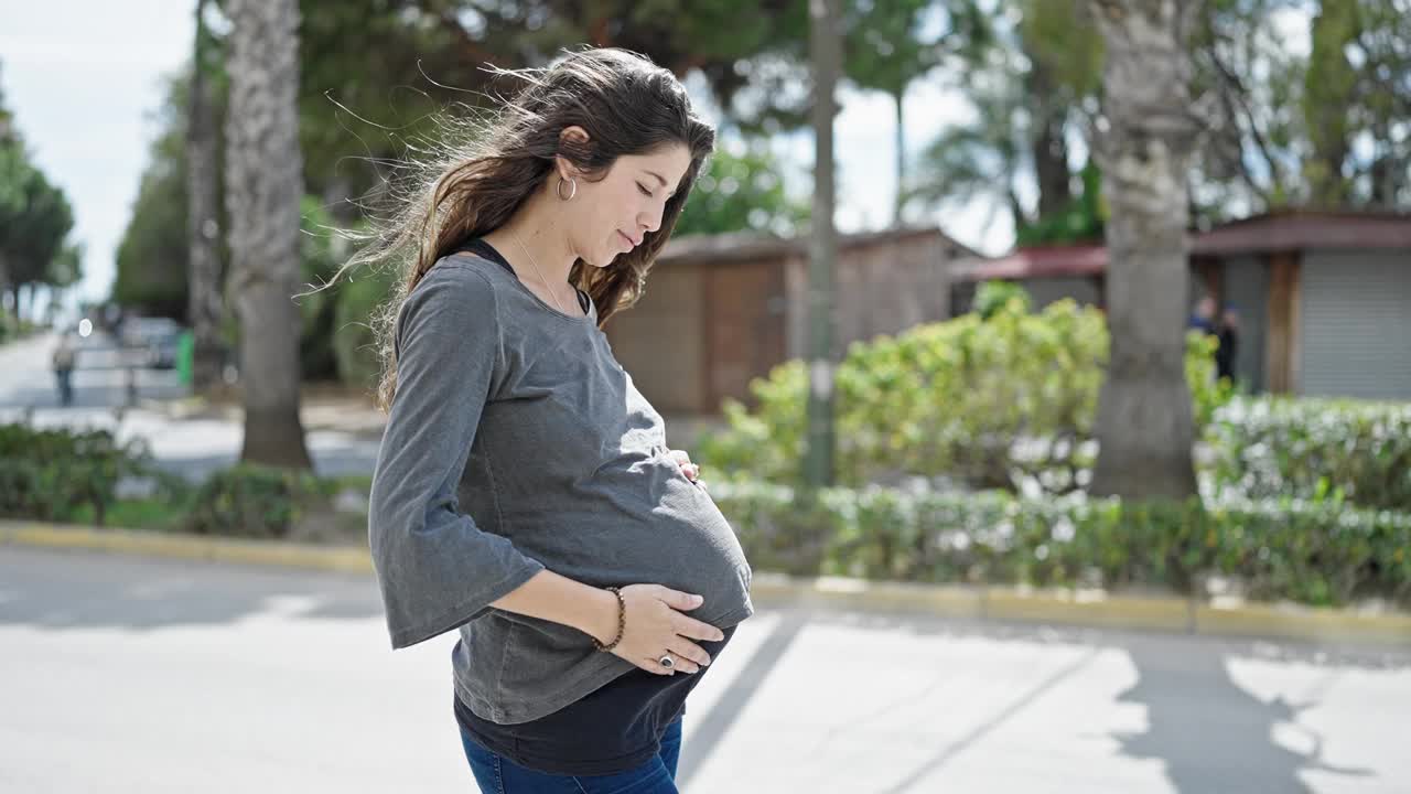 年轻的孕妇微笑着，自信地抚摸着公园里的肚子视频素材