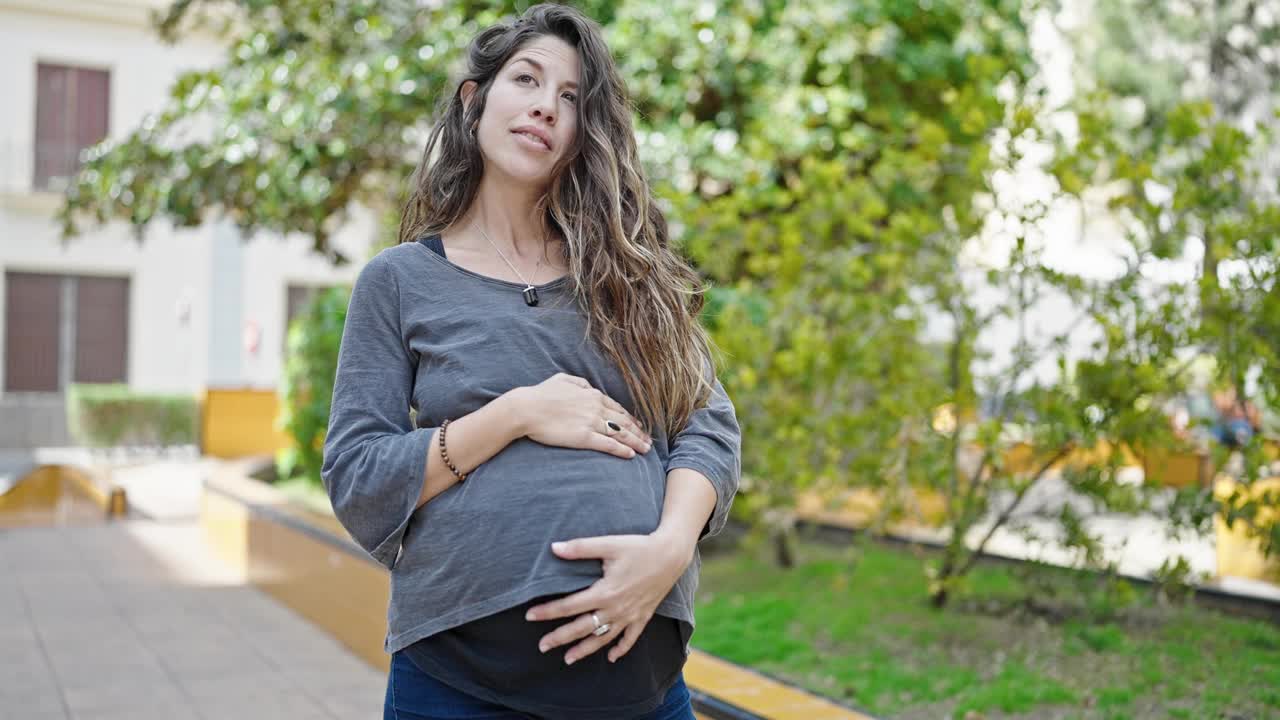 年轻的孕妇微笑着，自信地抚摸着公园里的肚子视频素材