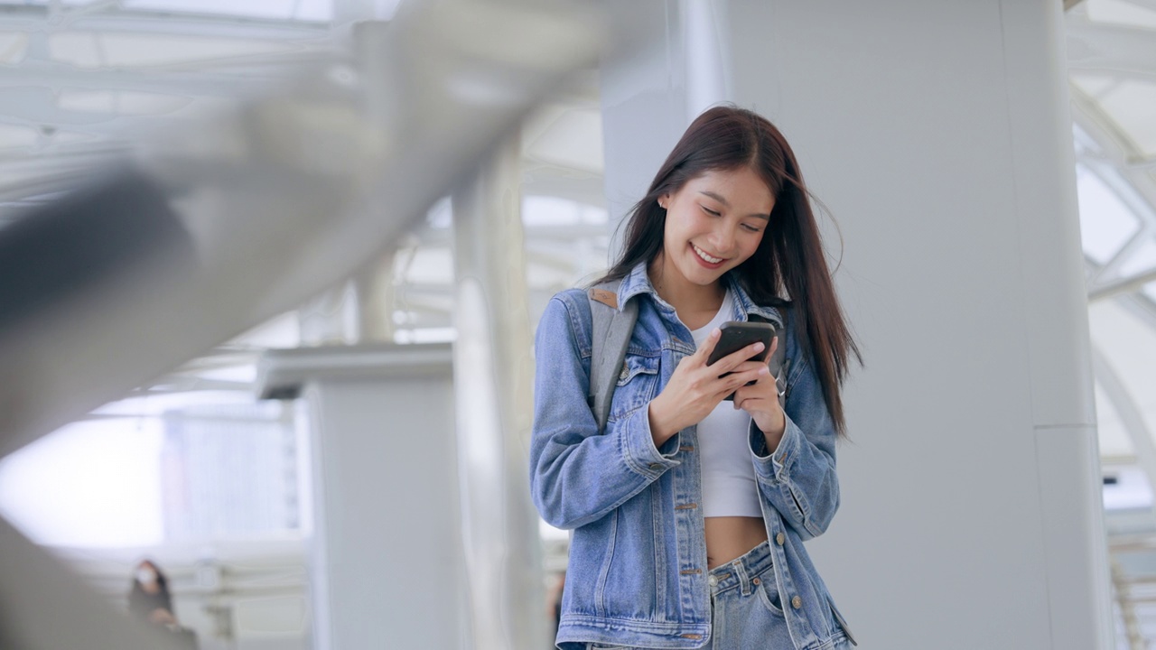 年轻的亚洲女子在户外微笑着使用手机。快乐的女游客穿着牛仔裤夹克，拿着智能手机出现在公共场合视频素材