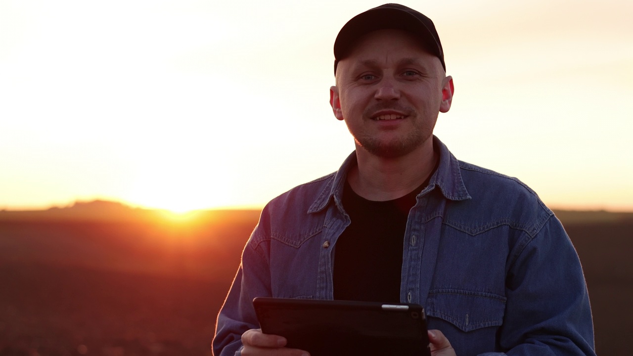 夕阳下，一位微笑着的男性农民在犁地的田地里拿着数码平板电脑，看着相机。检查和控制土壤质量，土地准备播种作物。智能农业技术视频素材