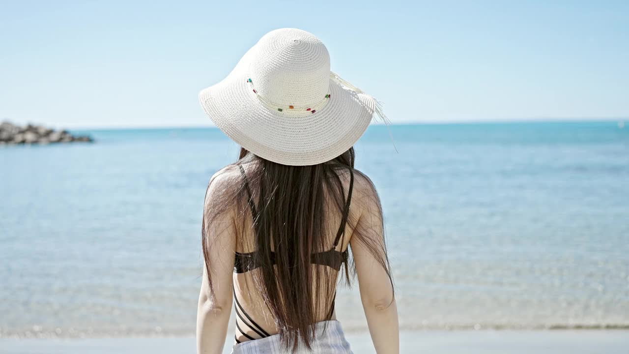 年轻的中国女游客穿着泳装，戴着夏帽，在海边自信地微笑着视频素材