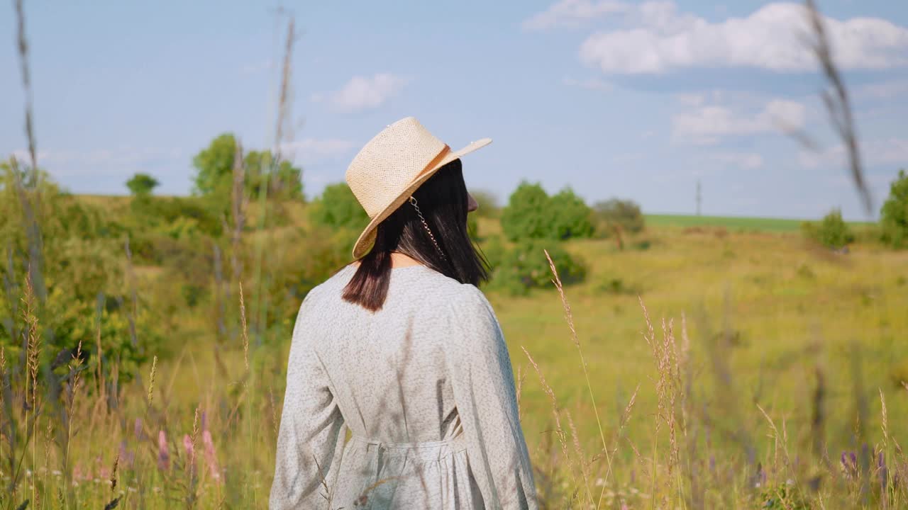 一个微笑的迷人的年轻成年女子在一个阳光明媚的夏天，穿着裙子和帽子在野外玩耍视频素材