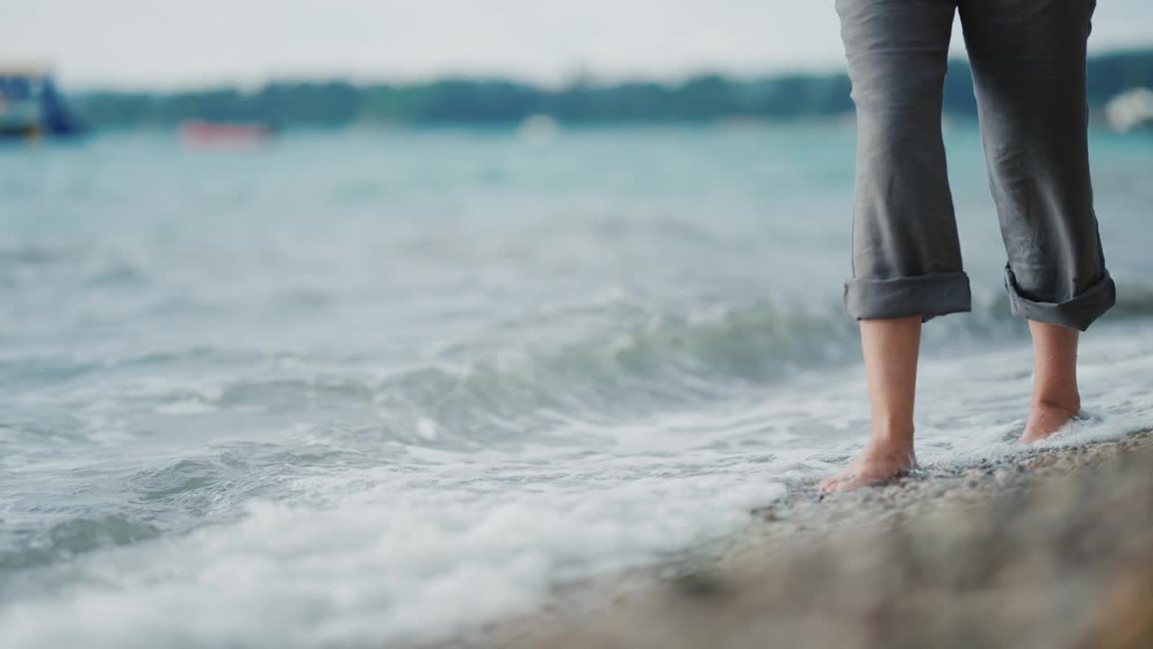 细细的女性腿和脚穿着裤子在沙滩上沿着海水行走，缓慢地溅起水和泡沫视频素材