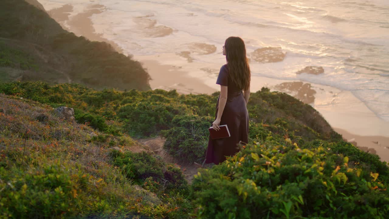 不知名的女人拿着书走在海边附近的山上。女人喜欢傍晚的海岸。视频素材