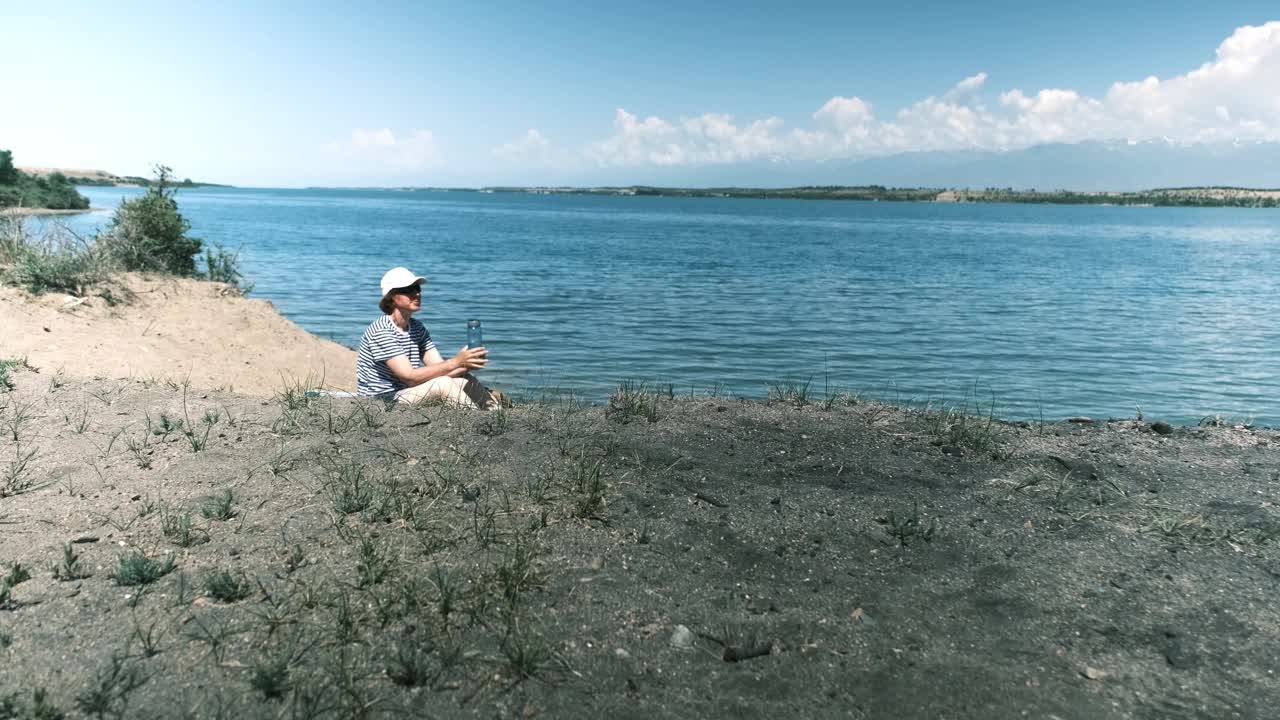 女游客坐在湖边沙滩上喝水视频素材