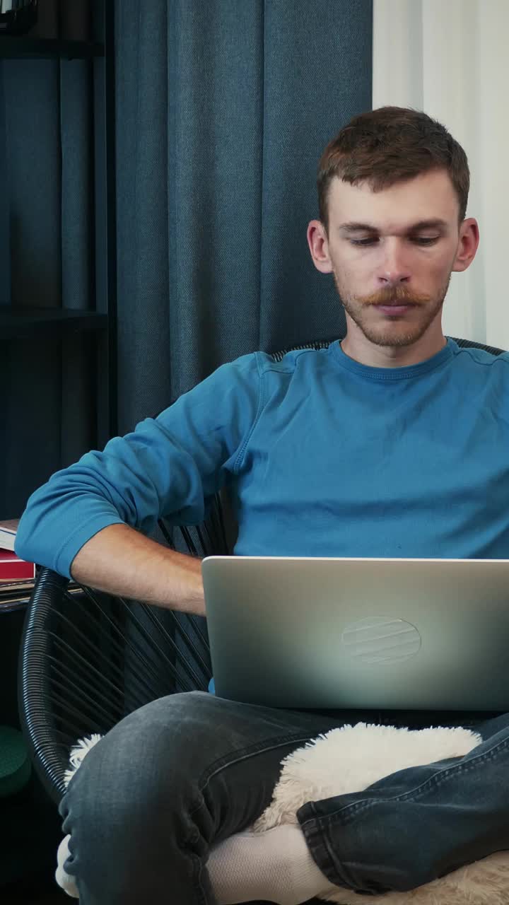 Man working on laptop while sitting on cozy couch in apartment. Vertical video视频素材