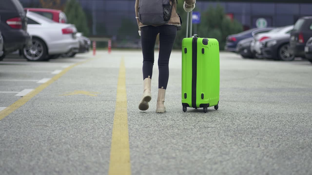 背着双肩包、梳着马尾辫的女旅行者推着大行李箱走在机场、火车站停车场，慢动作的电影镜头视频素材