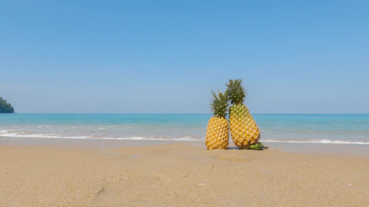 两个菠萝在蓝天碧海的美丽沙滩上。激起泡沫的海浪拍打着沙滩。在一个阳光明媚的夏日去天堂度假。阳光洒在沙滩上。视频素材