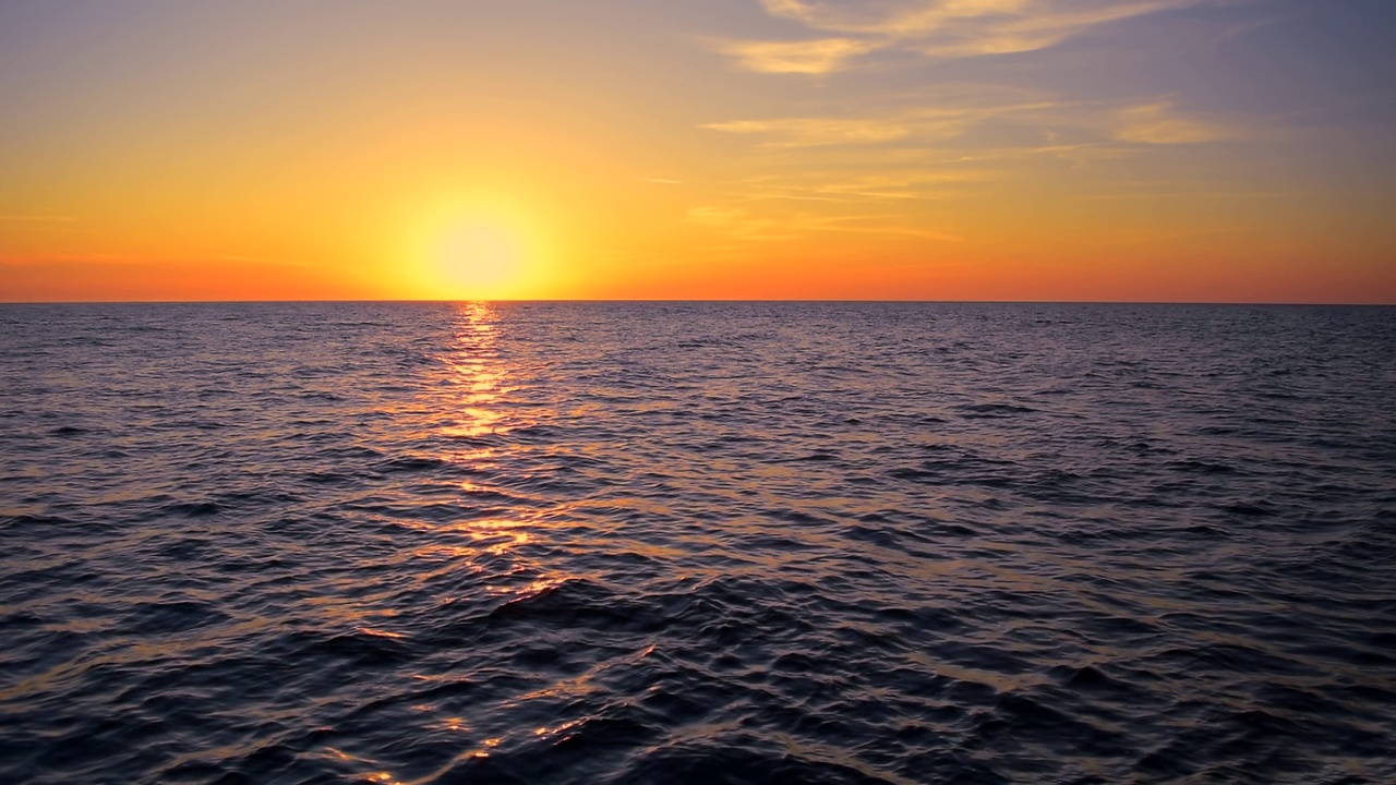 海浪反射黄色和金色的阳光在橙色和蓝色的日落背景。美丽的阳光与橙色发光的云在天空，平静的地平线场景由阳光明媚的海滩，中景，慢动作，高清。ProRes 422 HQ视频素材