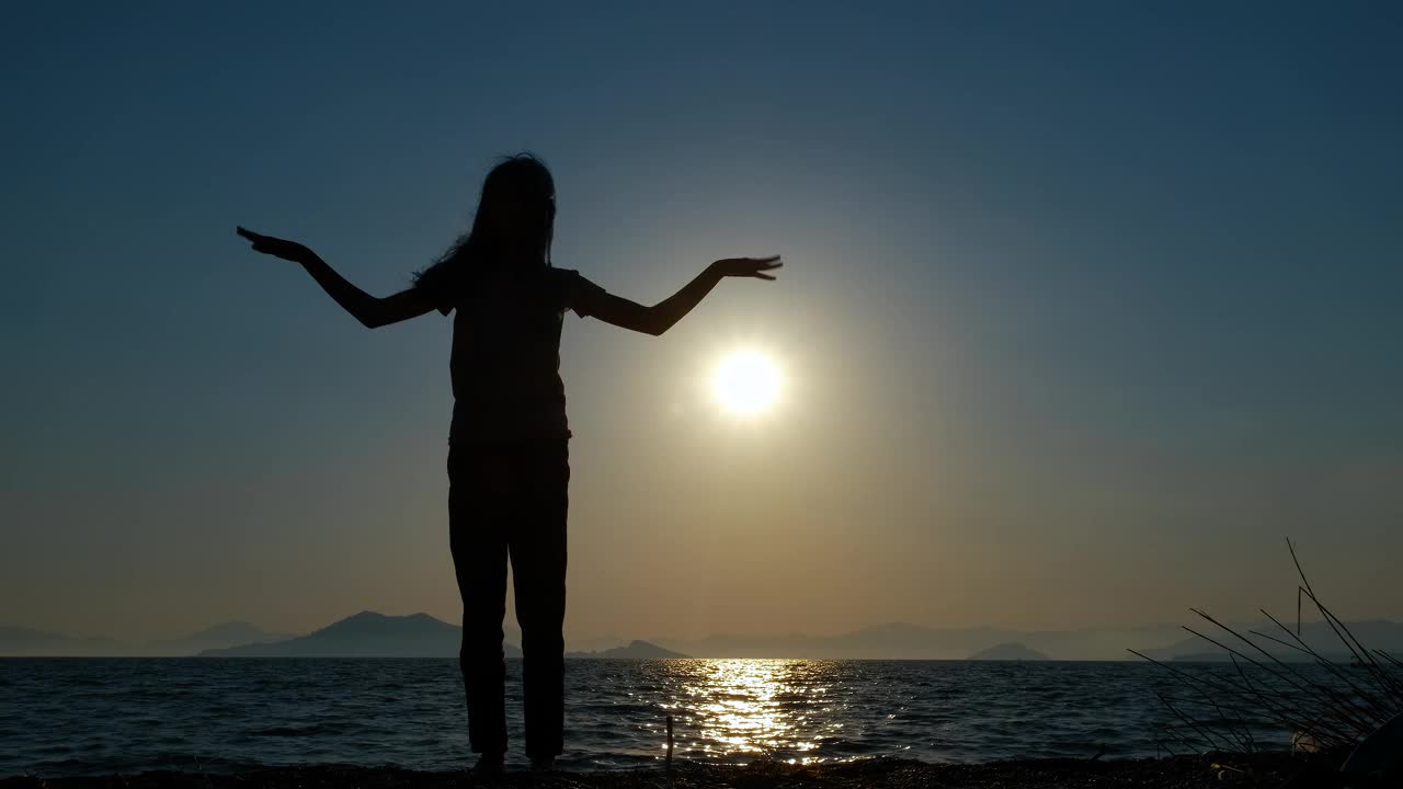 女孩的剪影在海湾上挥手。视频素材