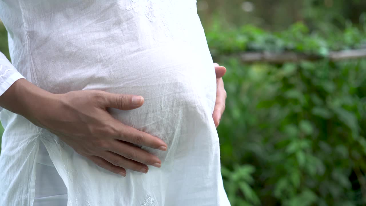 公园里身穿白衣的孕妇双手放在肚子上。怀孕，生育，准备和期望的概念。视频素材