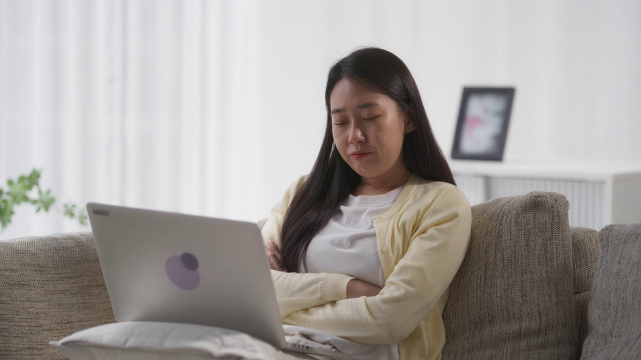 年轻美丽的亚洲女性浏览互联网，使用笔记本电脑在家里，计划一个令人兴奋的旅行行程，她的度假之旅，而坐在客厅的沙发上视频素材