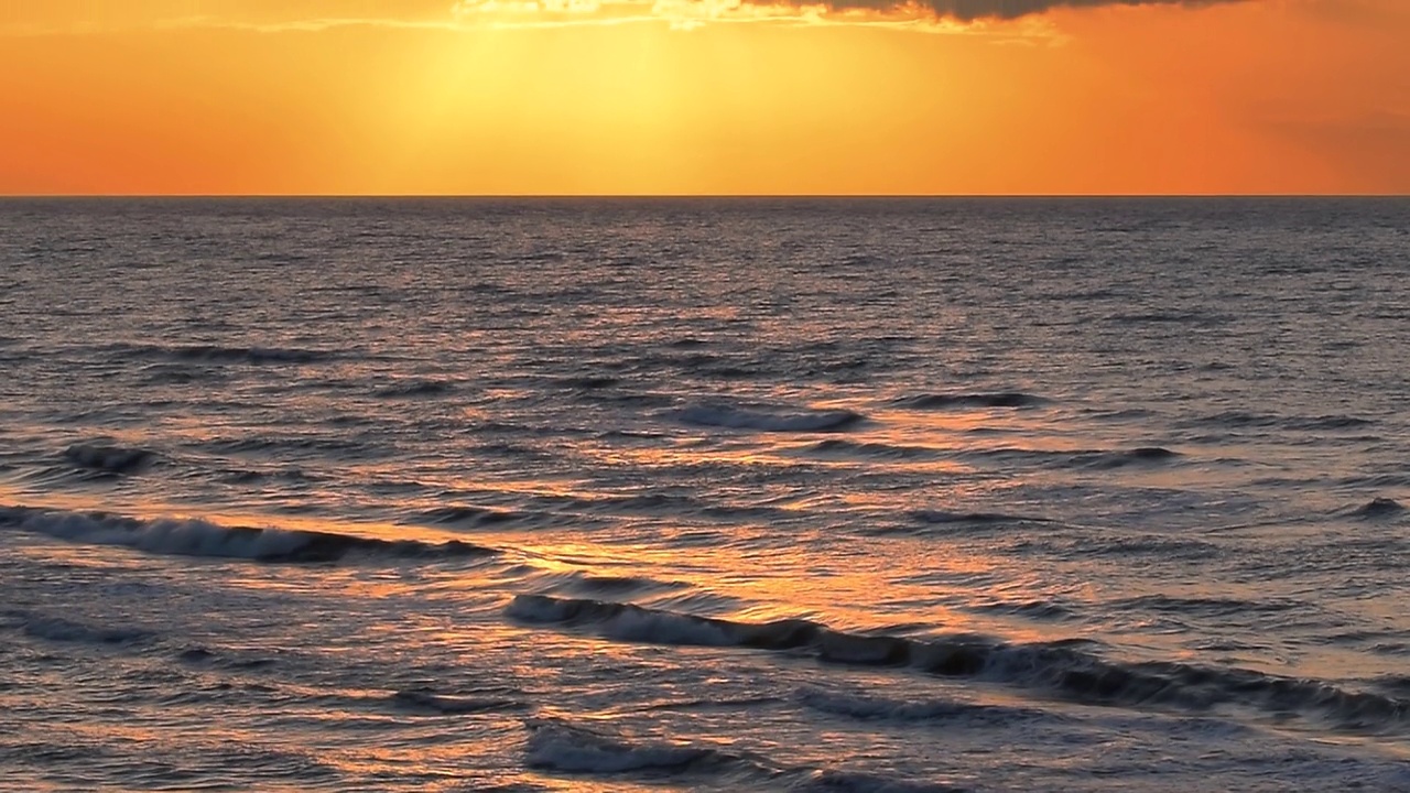 美丽的海浪反射着黄色和橙色的夕阳光芒，以海滩上五颜六色的天空云彩为背景，平静的地平线场景，中景，慢镜头。ProRes 422 HQ视频素材