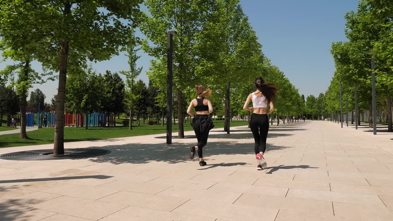 两个苗条的年轻女子在夏天跑过一座现代化的公园。运动。视频素材