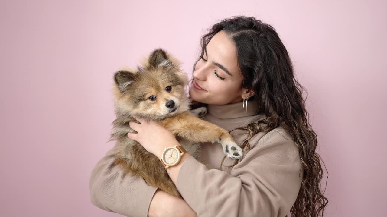 年轻的西班牙妇女带着狗微笑自信地站在孤立的粉红色背景上视频素材