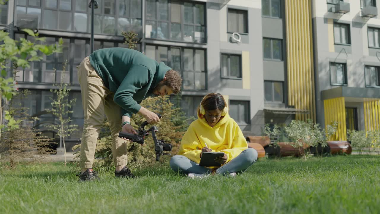 专业摄像师拿着相机在住宅区拍摄黑皮肤的女孩坐在草坪上，戴着耳机和平板电脑。后台。慢动作视频素材