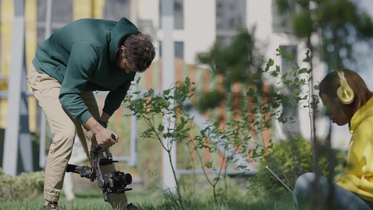 一名年轻的男摄影师在视频中拍摄了一个皮肤黝黑的女孩，她坐在一个住宅区的草地上的树荫下。后台。慢动作视频下载