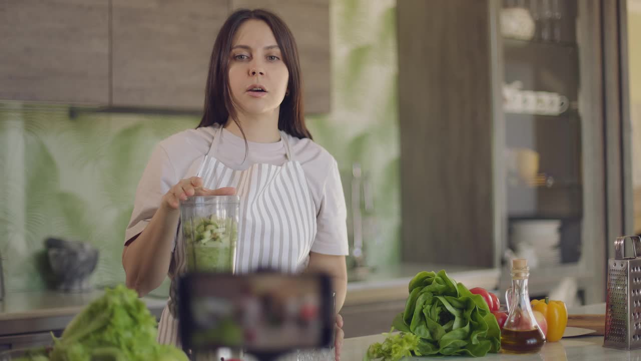 快乐的博主女孩谈论健康的生活方式和拍摄视频食谱。女孩对着智能手机摄像头看了看并说话。慢动作视频素材