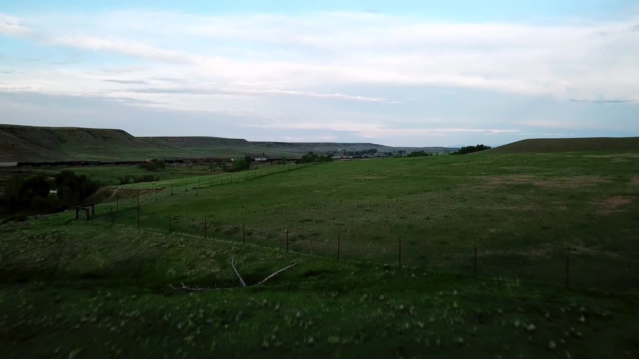 在蒙大拿州比林斯的黄昏，航拍的绿色山丘上，火车和河流映衬着天空，无人机向前飞行视频素材